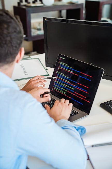 A person wearing a light blue shirt is seated at a desk, working on a laptop with a black screen displaying colorful lines of computer code. The individual's right hand is holding a mouse while their