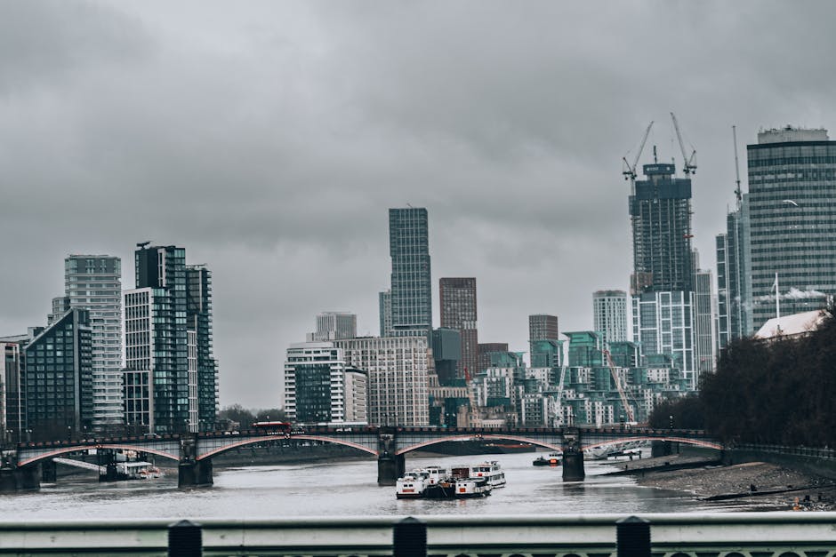 An aerial view of central London showing the River Thames curving through the cityscape. Prominent modern office buildings with glass facades, some with rooftop terraces, are located along the riverba